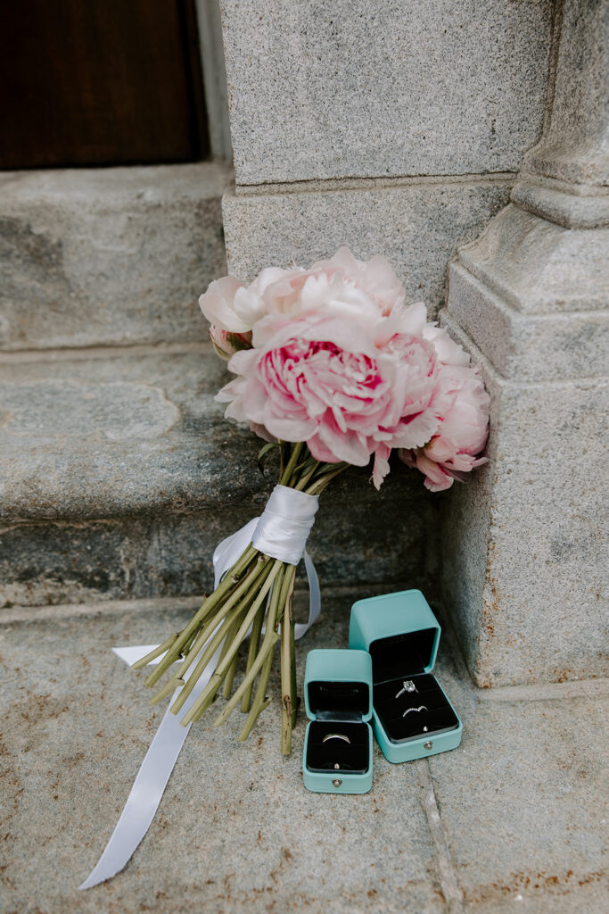 Bouquet of soft pink peonies tied with white ribbon resting on stone steps beside two open blue ring boxes holding the wedding bands and engagement ring, Cambridge City Hall wedding details photographed by Boston elopement photographer Genevieve Photography