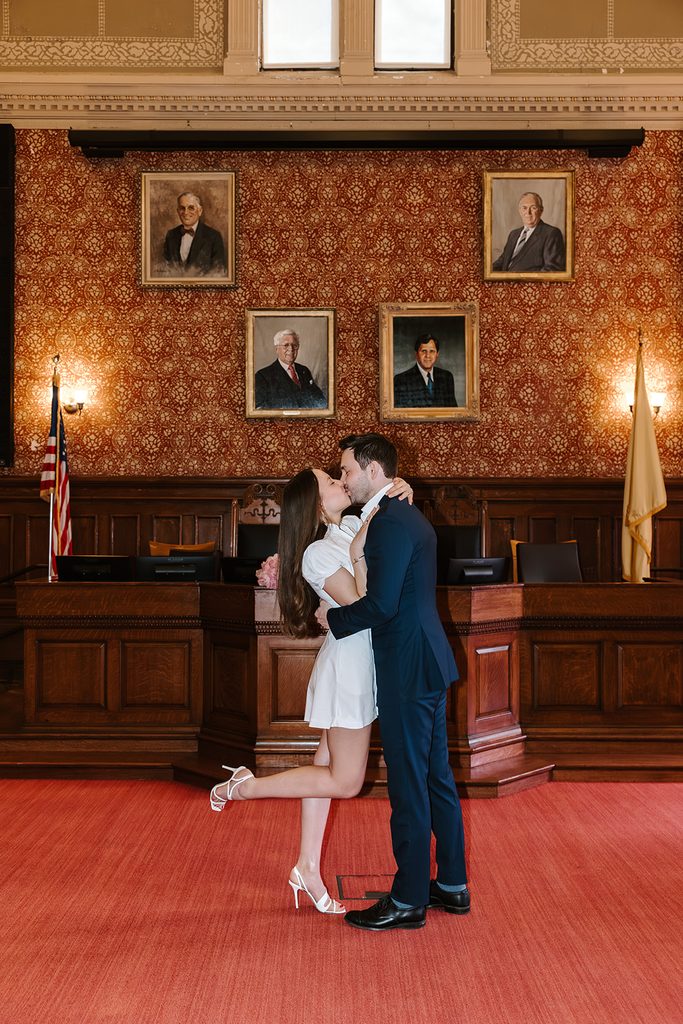 Bride kicking up her heel as she kisses her groom inside the historic council chamber at Cambridge City Hall, with red patterned walls, framed portraits, and warm wall sconces behind them. Cambridge City Hall wedding photographed by Boston elopement photographer Genevieve Photography