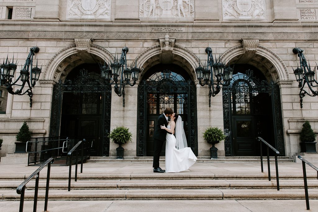 Bride and groom kissing on the front steps of the Boston Public Library McKim Building in Copley Square, with iron lanterns and arched doorways behind them and her veil blowing in the breeze. Boston elopement photographed by Boston elopement photographer Genevieve Photography