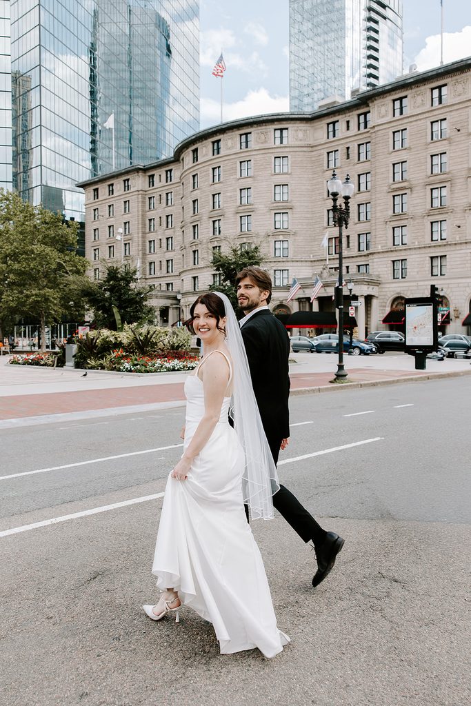 Bride smiling back at the camera as she walks with her groom across the street in Copley Square, with the Fairmont Copley Plaza hotel and glass towers behind them, after their Boston Public Library elopement. Photographed by Boston elopement photographer Genevieve Photography