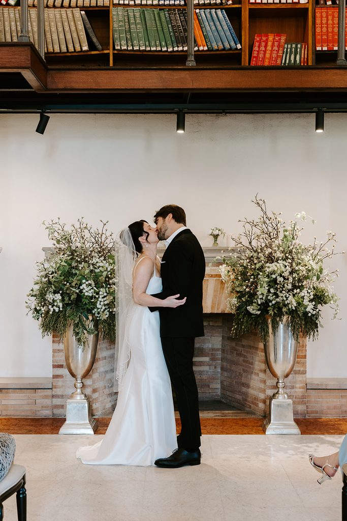 Bride and groom sharing their first kiss in front of the fireplace in the Guastavino Room at the Boston Public Library, with large white floral arrangements in silver urns on each side and colorful books above. Boston Public Library wedding photographed by Boston elopement photographer Genevieve Photography